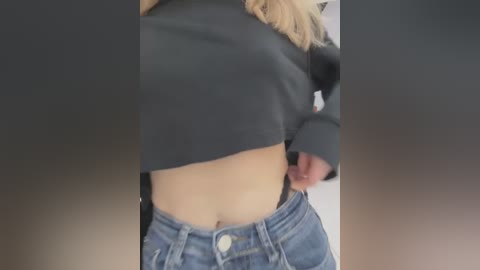 Video of a woman's torso, midsection to waist, wearing a black cropped top and high-waisted blue jeans, blonde hair visible. Background is blurred, neutral colors.