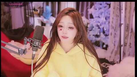 Media: A video of a young Asian woman with long brown hair, wearing a yellow hoodie, singing into a microphone in a cozy room with floral wallpaper.