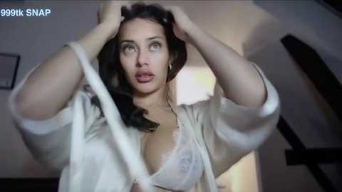 Media: Video of a woman in a white blouse, with her hands in her hair, looking surprised. Background shows a dimly lit room with a staircase.