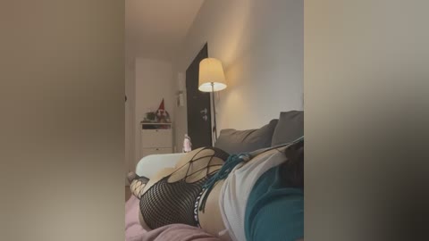 Media: Video of a cozy bedroom with a beige wall and a bed with a white duvet and a teal blanket, featuring a person wearing a black fishnet top and blue jeans. A floor lamp with a white shade casts a soft glow.