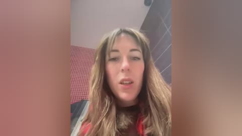 Media: Video of a Caucasian woman with long, wavy brown hair and fair skin, wearing a red shirt, in a bathroom with red and grey tiled walls.