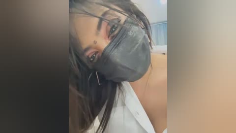 Media: A close-up video of a woman with long, dark hair and light skin, wearing a black face mask, white shirt, and a small nose piercing, in a blurred, indoor setting.