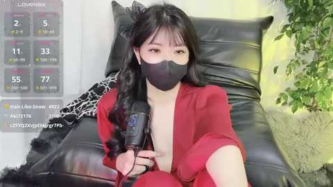 Media: Video of a young Asian woman with long black hair, wearing a black face mask, red suit, and black feather boa, sitting on a black leather chair, holding a microphone, in a modern indoor setting.