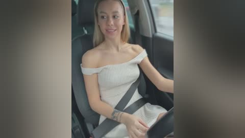 Media: Video of a smiling, fair-skinned woman with long blonde hair, wearing a white off-shoulder dress and seatbelt, sitting in a car, blurred background.