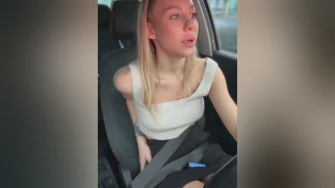 Video of a blonde, fair-skinned woman in a white off-shoulder top and black pants, sitting in a car, looking surprised.