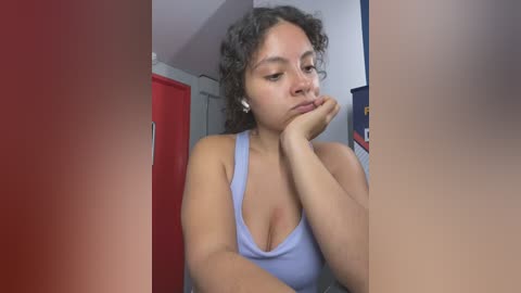 Media: Video of a young woman with curly hair, wearing a light blue tank top, resting her chin on her hand, in a dimly lit room with red and blue cabinets.