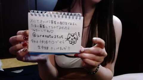 Media: Video of a young woman with long dark hair, holding a handwritten sign with Japanese text, wearing a white top. Background is dimly lit, showing a room with dark furniture.