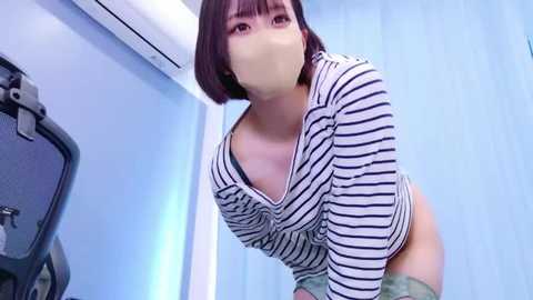 Media: Video of an Asian woman with short brown hair, wearing a black-and-white striped top and mask, bending over in a bathroom with light blue walls and an air conditioner.