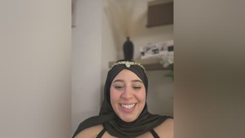 Media: Video of a smiling, dark-skinned woman with a medium build, wearing a black hijab adorned with a gold brooch and a black top, seated in a beige room with a vase of dried flowers on a shelf.