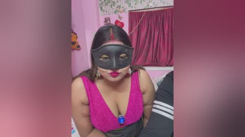 Media: A video of a woman with medium skin tone and dark hair, wearing a black Venetian mask, bright pink dress, and large hoop earrings, seated indoors with pink walls, floral wallpaper, and a red curtain.