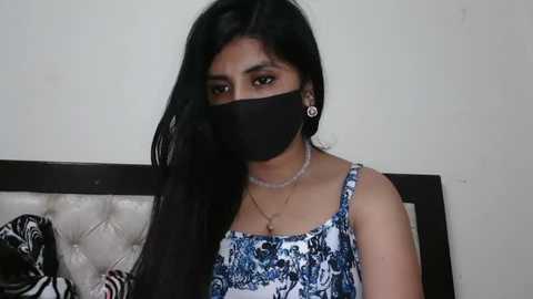 Media: A video of a South Asian woman with long black hair, wearing a black mask, blue-patterned sleeveless top, and silver earrings, seated on a tufted, black leather chair with a floral pattern.