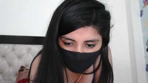 Media: A video of a young woman with long, straight black hair, wearing a black mask and a red top, leaning forward, with a white tufted headboard in the background.