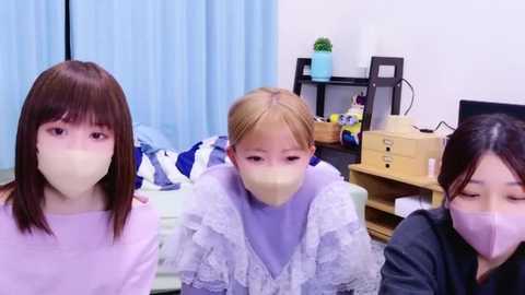 Video of three young women, two with light skin and straight brown hair, one with light skin and blonde hair, wearing surgical masks, seated indoors in a cluttered room with blue curtains, wooden shelves, and a TV.