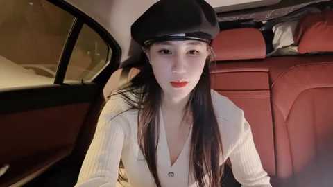 Media: Video of a young Asian woman with long black hair, wearing a black beret, light-colored cardigan, and red lipstick, seated in a red-leather interior of a car.