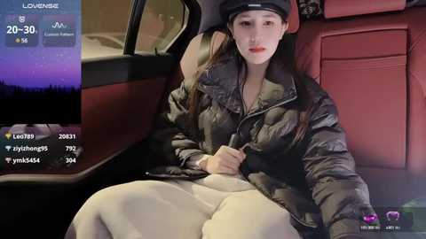 Media: A video of a young woman with long hair, wearing a black puffer jacket, beige pants, and a black cap, sitting in a red leather car seat, looking relaxed.