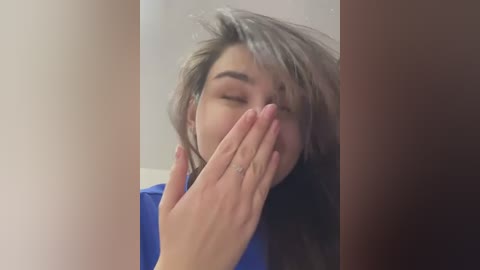 Media: A video of a young woman with long, tousled blonde hair covering her eyes, covering her mouth with her hand, wearing a blue shirt, taken in a dimly lit room with beige walls.