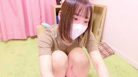 Media: Video of an Asian woman with straight, shoulder-length brown hair, wearing a beige sweater and white face mask, kneeling on a green carpet in a bedroom with pink curtains and wooden furniture.