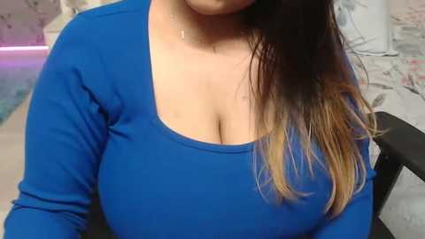 Media: A video of a woman with long, straight, dark hair with light blonde ends, wearing a tight-fitting, bright blue long-sleeve shirt that accentuates her large breasts. The background is a blurry, indoor setting with a light-colored bed and floral-patterned bedding.