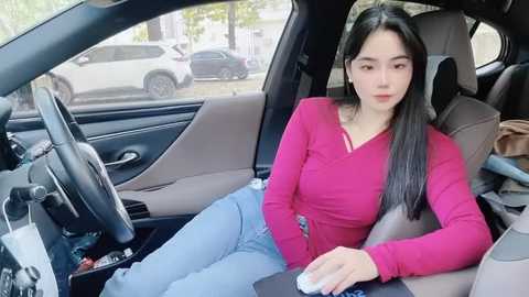 Media: Video of an Asian woman with long black hair, wearing a pink long-sleeve top and blue jeans, seated in a modern car, holding a white mouse, with a blurred street and parked cars visible through the window.