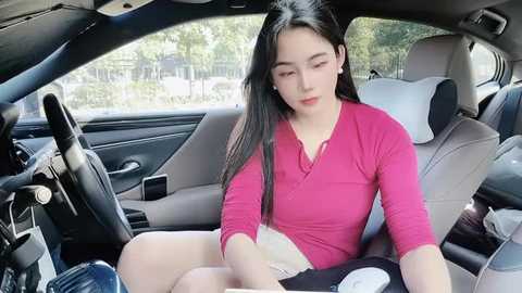 Media: Video of an East Asian woman with long black hair, wearing a pink long-sleeve top and beige shorts, sitting in a car, looking down at her phone, with a blurred, green background.