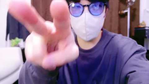 Media: A video of an Asian man with glasses, mask, and dark hair, wearing a gray shirt, making a peace sign with his right hand in a dimly lit room.