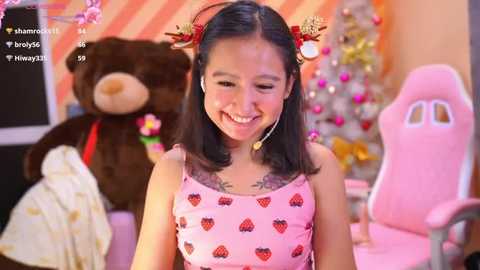 Media: A video of an Asian woman with black hair, wearing a pink strawberry-patterned dress, smiling, standing in a cozy, festive room with a Christmas tree and plush toys.