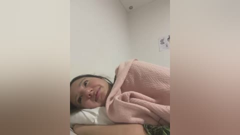 Media: A video of an Asian woman with dark hair and light skin, lying in a bed with a pink quilt, in a small, dimly lit room with white walls and a poster on the wall.