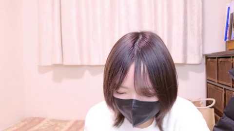 Media: A video of a young Asian woman with straight, shoulder-length dark hair, wearing a black face mask, white shirt, and sitting on a wooden floor. Background includes a beige curtain, a wooden cabinet, and a laundry basket.