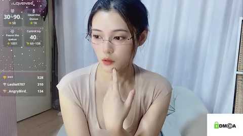 Media: A young East Asian woman with fair skin and black hair, wearing glasses, sits in a beige top, looking contemplative in a dimly-lit room with white curtains.