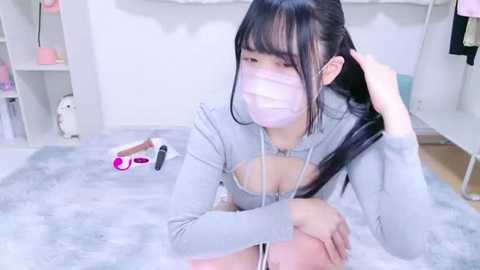 Media: Video of an East Asian woman with long black hair, wearing a gray hoodie and a white face mask, sitting on a plush gray carpet, holding a pink vibrator. The background features a white room with shelves and a teddy bear.