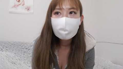 Media: Video of an Asian woman with long brown hair, wearing a white surgical mask, gray hoodie, and a gray bedspread with white fur trim.