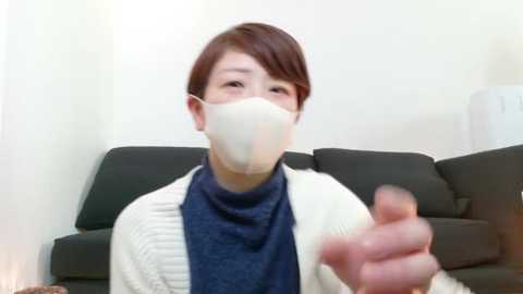 Media: Video of an East Asian woman with short brown hair, wearing a white face mask, blue sweater, and white cardigan, giving a thumbs-up gesture, seated on a black couch in a minimalist living room.