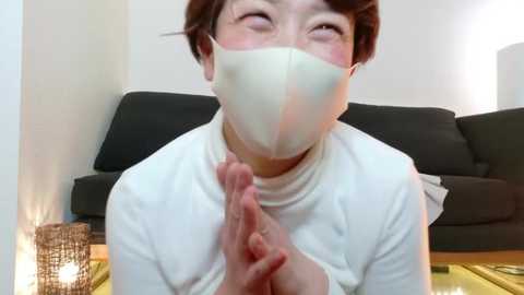 Media: Video of an Asian woman with short brown hair and a white face mask, wearing a white long-sleeve shirt, clasping her hands in prayer, in a minimalist living room with a dark sofa, white walls, and a lit candle.