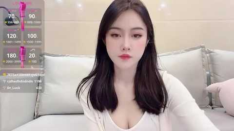 Media: Video of an East Asian woman with long black hair, wearing a white top, sitting on a white couch. Background shows a minimalist, modern room with beige walls and soft lighting.