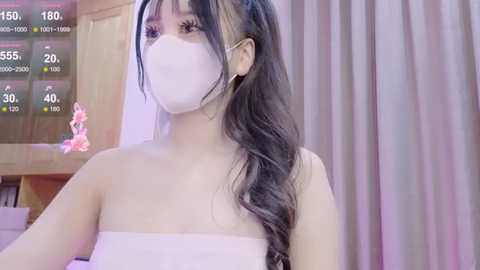 Media: Video of an Asian woman with long, wavy black hair, wearing a white face mask and a strapless white towel, indoors with a wooden door and a digital clock showing 150%.