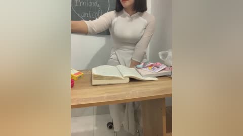 Media: Video of a woman with fair skin, shoulder-length dark hair, wearing a white dress, sitting at a wooden desk in a classroom. She writes on a chalkboard behind her. Desk has books, a pen, and a toy car.