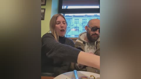 Media: Video of two people, a blonde woman with a dark sweater and a bald man with glasses, eating in a dimly lit restaurant with a large window showing a city scene.