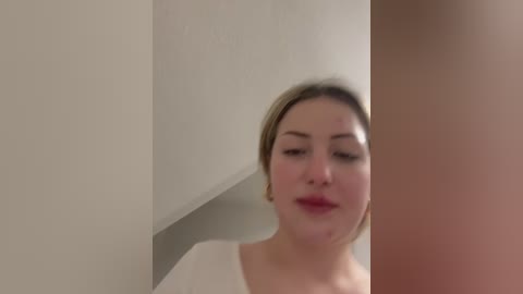 Media: A video of a young Caucasian woman with light skin and blonde hair, wearing a white top, looking into a mirror. The background is plain, off-white.