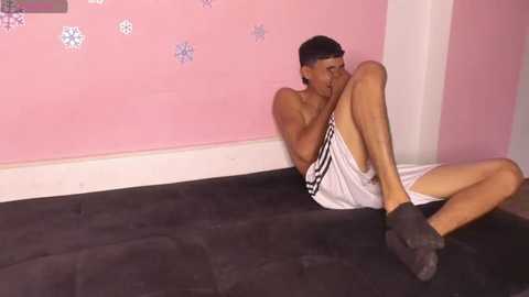 Media: Video of a shirtless, slender, dark-skinned man with short black hair, wearing white shorts, sitting on a black mat, hiding his face with his hands, in a pink room with white walls and snowflake decals.