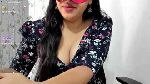 Media: Video of a plus-sized woman with long black hair, wearing red sunglasses, a low-cut floral dress, and dangling earrings, sitting with arms crossed. Background features floral wallpaper.