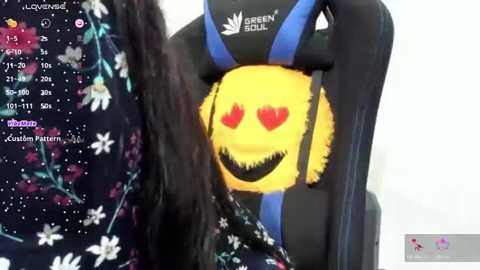 Media: Video of a woman with long, dark hair wearing a black and blue gaming chair with a smiling yellow face emoji featuring red hearts as eyes. Background includes a colorful digital wallpaper.