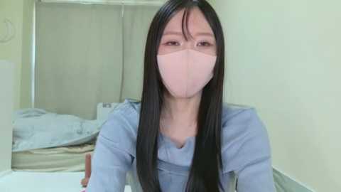 Media: Video of a young Asian woman with long black hair, wearing a light blue nurse's uniform and a face mask, standing in a hospital room with beige curtains and a bed in the background.