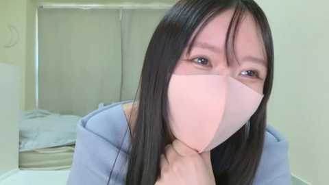 Media: Video of a young Asian woman with long black hair, wearing a pink face mask and a blue hoodie, sitting indoors with cream-colored walls and a bed visible in the background.