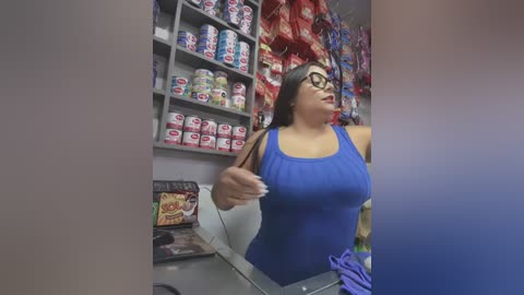 Media: Video of a plus-sized woman in a blue tank top, glasses, and long brown hair, gesturing in a brightly lit candy store with colorful shelves filled with assorted candies.