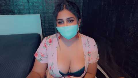 Media: Video of a woman with medium skin tone, wearing a floral pink blouse and a teal face mask, revealing cleavage, sitting on a black bed. Background shows a dark curtain and a white headboard.