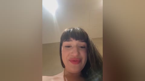 Media: Video of a smiling, light-skinned woman with straight, dark brown hair and bangs, standing in a beige-tiled bathroom with a white sink and mirror in the background.