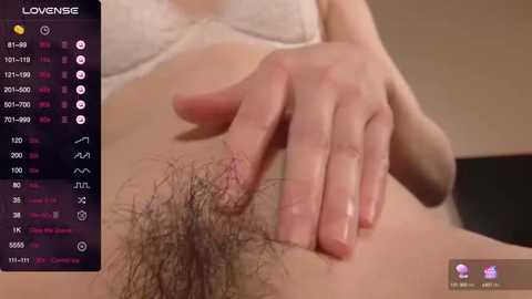 Media: Close-up video of a woman's lower torso, showing a hand with manicured nails gently touching her pubic hair, which is thick and dark. She wears a white bra. The background is blurred.