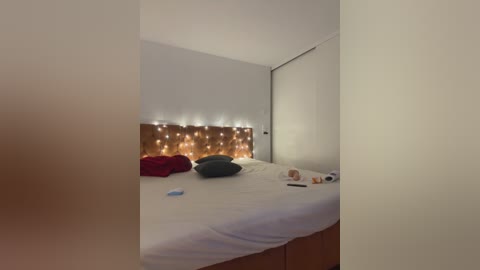 Media: Video of a minimalist bedroom with a neatly made bed, white linens, and a cozy, warm LED-lit wooden headboard. A red pillow and a grey one sit on the bed. The room has soft, ambient lighting.