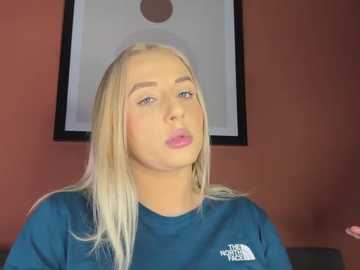 Video of a blonde, fair-skinned young woman with pink lipstick, wearing a blue shirt, in a room with a framed picture on a red wall.