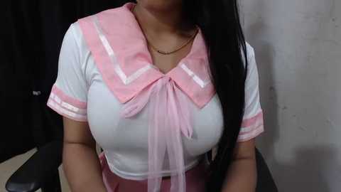 Media: Video of a woman with long black hair, wearing a white blouse with pink accents, sitting against a beige wall, wearing a gold necklace.